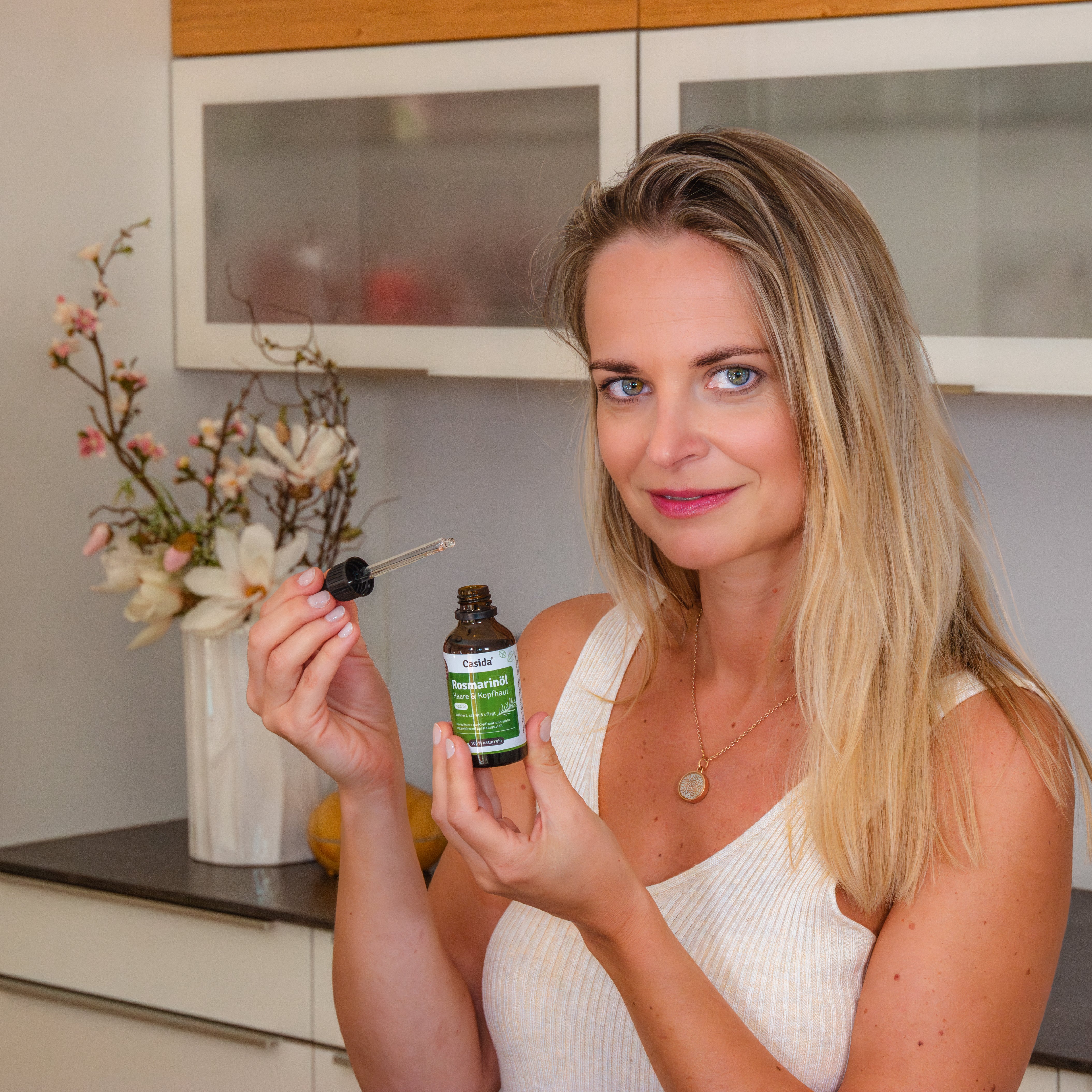 Rosmarinöl for hair and scalp in a dark glass bottle, held by a woman with long blonde hair, in a modern kitchen setting.