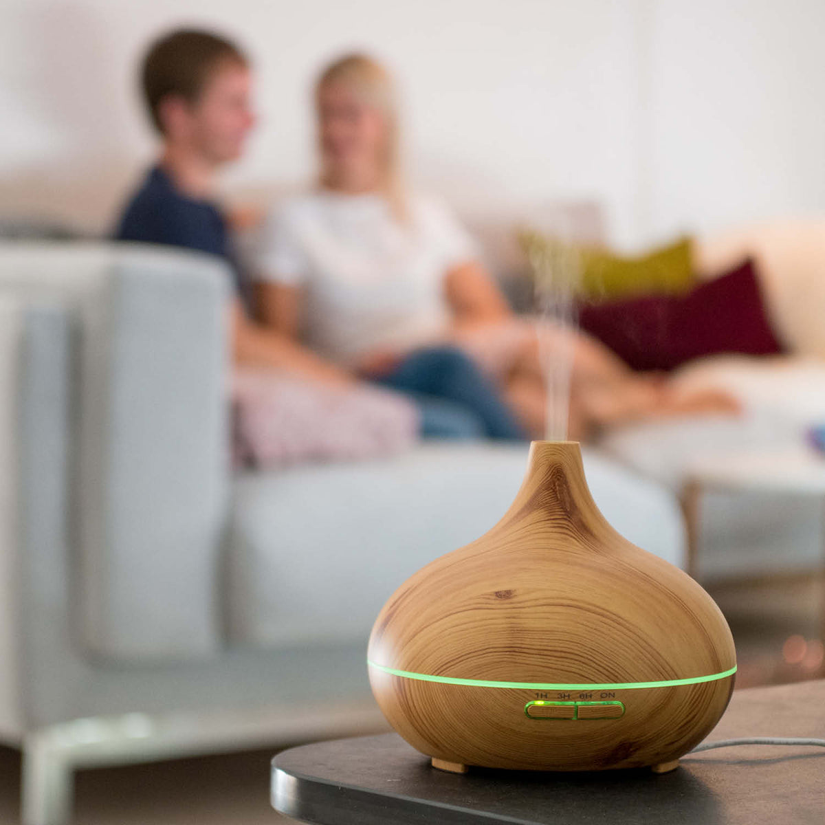 Aroma diffuser in wood design with LED lighting, emitting mist, placed on a table with a blurred background of two people.
