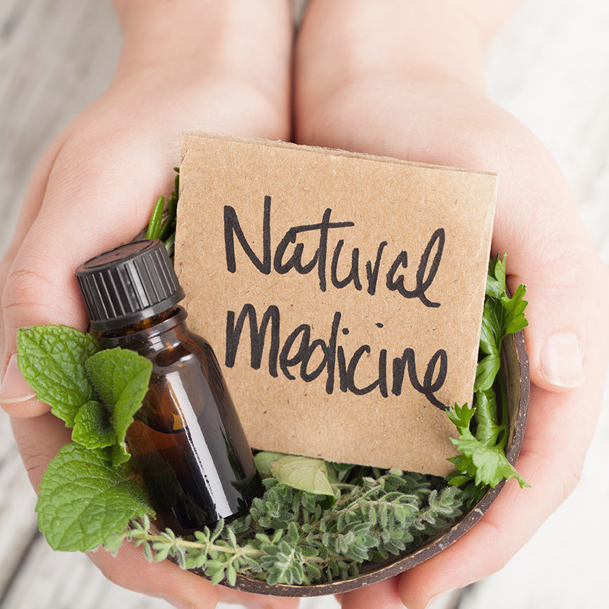 Zwei Hände halten eine Schale mit Kräutern einer kleinen gefüllten braunen Glasflasche und einem Schild auf dem Natural Medicine steht