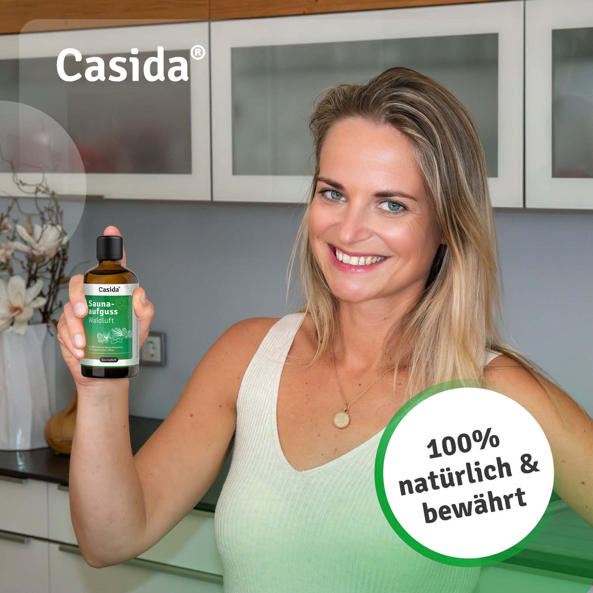 Casida saunaaufguss waldluft oil in a bottle held by a smiling woman in a kitchen setting.